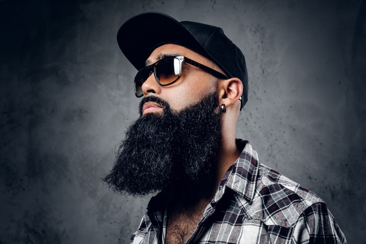 Pensive Black bearded male dressed in a fleece shirt, sunglasses and baseball cap posing over grey background.